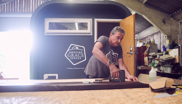 A person is sawing some wood. Behind him is a shipping container which has been adapted into housing. It is black with a white logo of 'Amazing Grace Spaces'