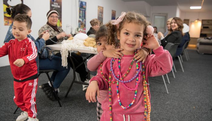 three children are at the fore front of the image wearing various necklaces. Behind them is a table with adult women taking part in knitting