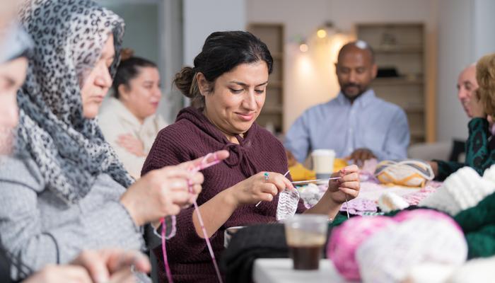 A group of women are knitting. The focus is a woman in the centre, who is looking down at her knitting. Behind them you can see two men and another lady, blurred.