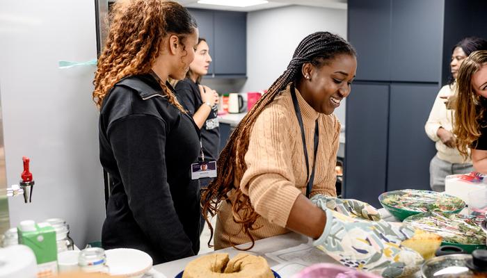 A group of women are standing behind a table laden with food, and serving. They all look happy and smiling