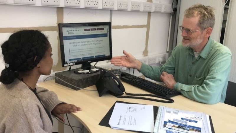 A man and a woman sit opposite each other, looking at a computer screen on the desk between them